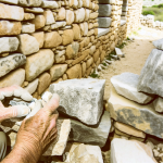 "A vintage pair of hands delicately working on a stone masonry restoration project of a heritage home, highlighting the intricate dry stone walling technique with different sizes and shapes of stones piled without the use of mortar."